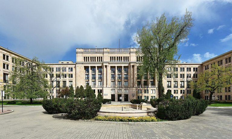 Widok na monumentalny, symetryczny dziedziniec gmachu Ministerstwa Finansów. Budynek ma jasną elewację z licznymi oknami i kolumnadą w części centralnej. Przed budynkiem znajduje się plac wyłożony kostką, niska zieleń w formie żywopłotów oraz pusta fontanna.
