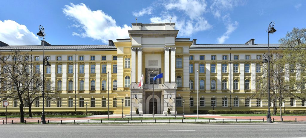 Szeroka panorama fasady klasycystycznego, żółto-białego gmachu Ministerstwa Finansów w Warszawie. Przed budynkiem znajduje się szeroki chodnik i ulica, a po bokach rosną drzewa. Nad centralnym wejściem z kolumnami powiewają flagi Polski i Unii Europejskiej.
