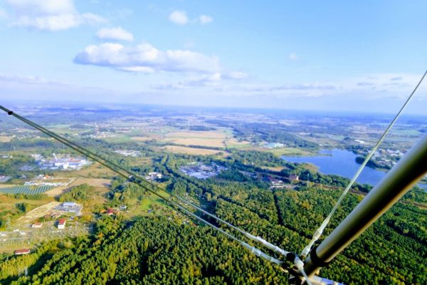 Widok z lotu ptaka, prawdopodobnie z paralotni lub motolotni (widoczne elementy konstrukcyjne i linki), na rozległy krajobraz wiejsko-leśny. Widać zielone lasy, pola uprawne, małe jezioro po prawej stronie oraz rozproszone zabudowania pod błękitnym niebem z lekkimi chmurami.